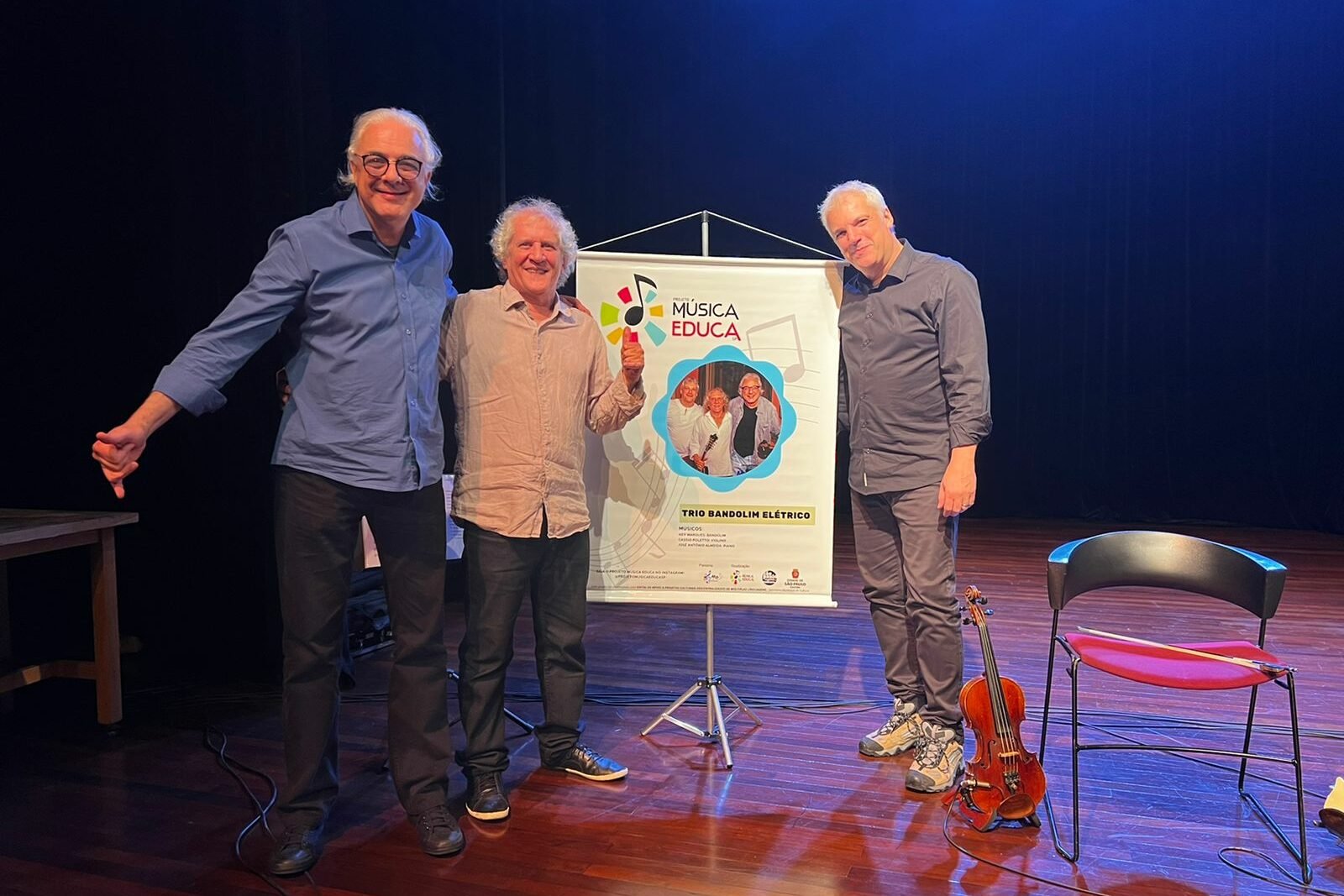 Ney Marques com os dois músicos do trio Bandolim Elétrico no palco com um banner do projeto Música Educa
